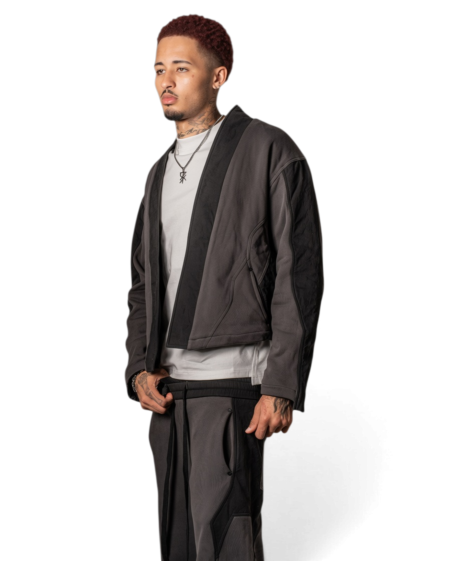 A man modeling a dark gray asymmetrical jacket with black trim, worn over a light gray t-shirt and dark gray pants, photographed against a solid white background.